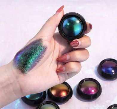 Fard à paupières caméléon en godets noirs, aux pigments irisés bleu, vert et violet, avec un swatch multicolore sur la main.